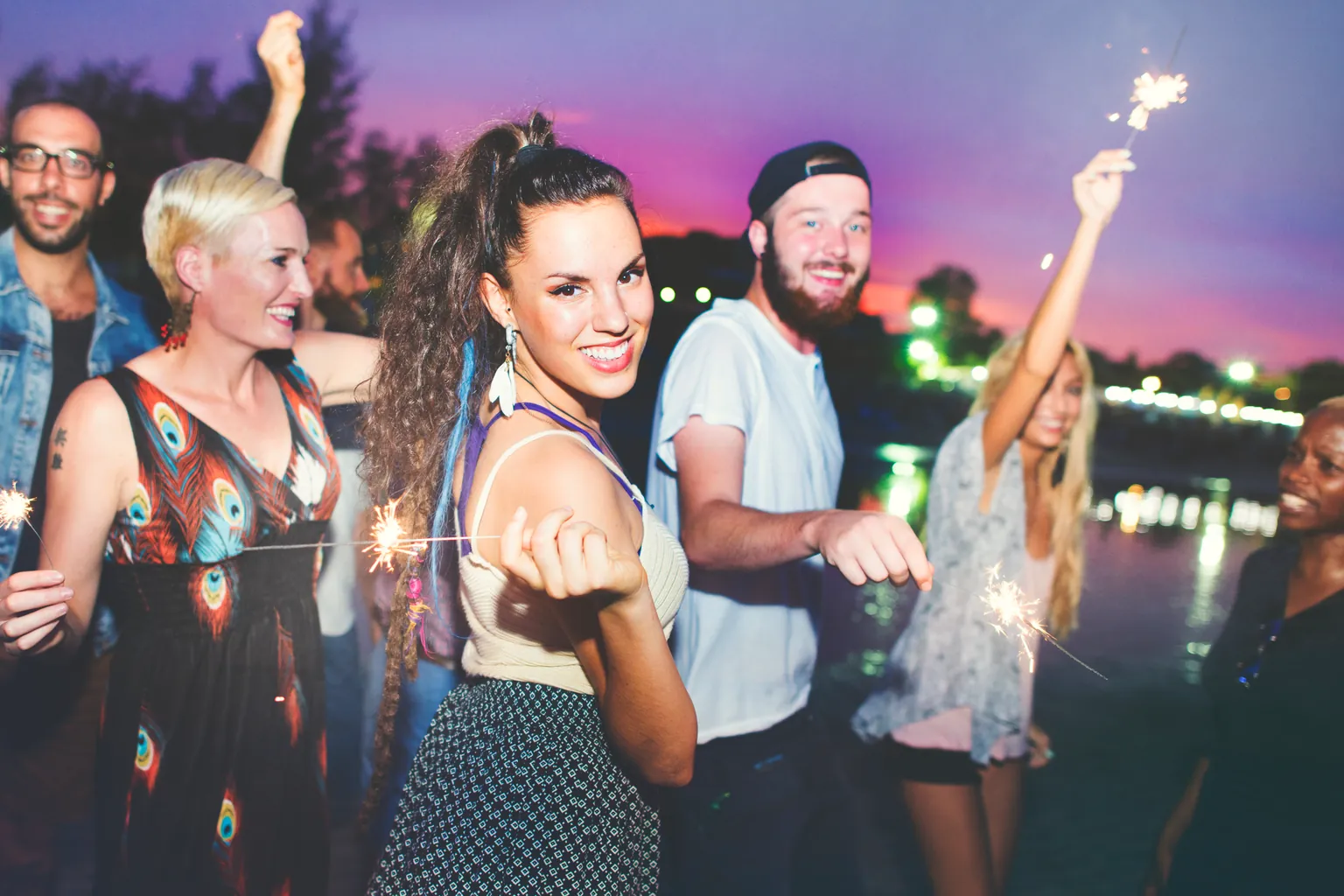 Friends stand by the water at dusk holding lit sparklers under a pink and purple sky