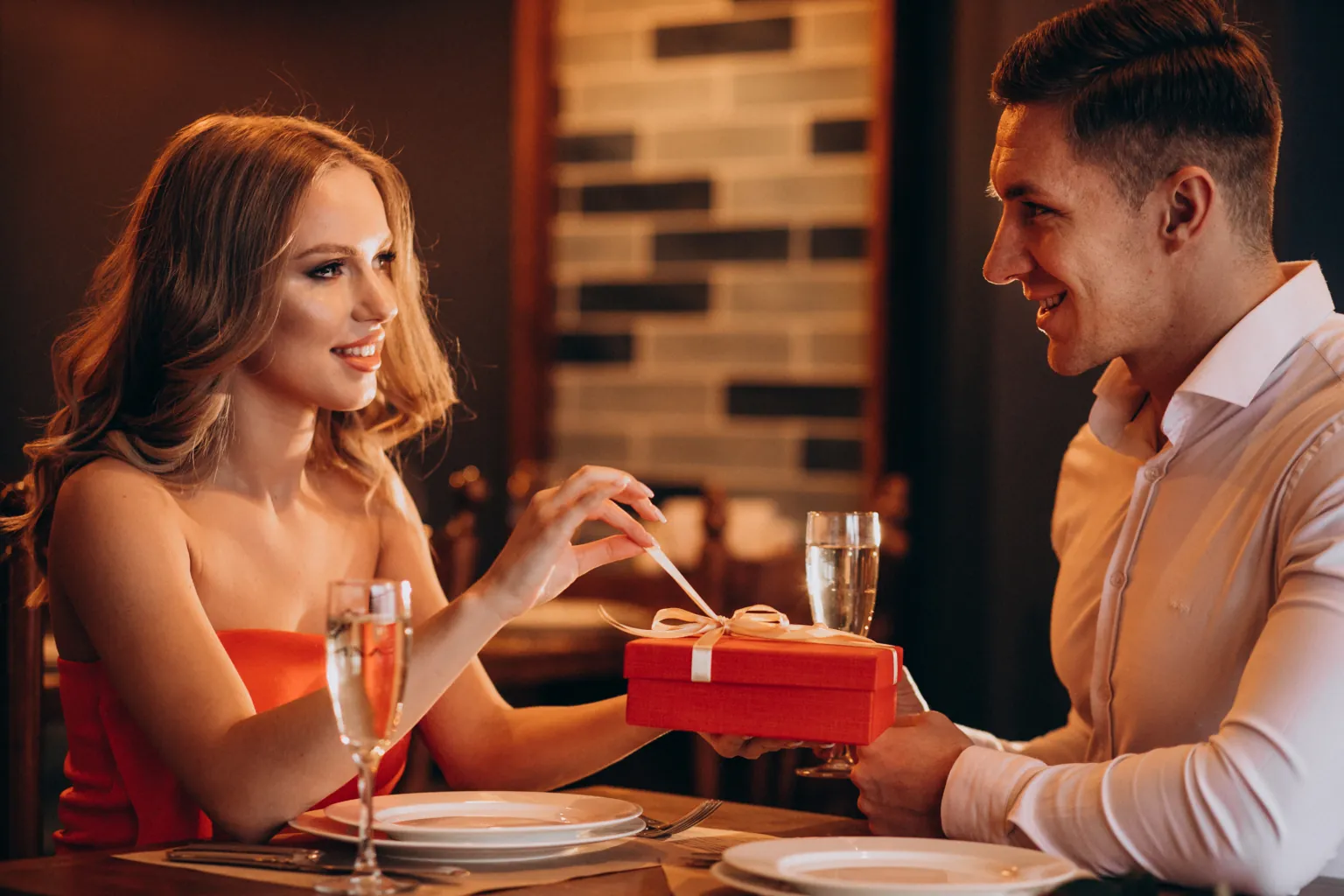 A man passes a red gift box tied with cream ribbon to a woman across a restaurant table in warm light