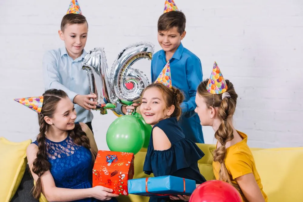 Five children in party hats hold presents and a silver foil balloon shaped as the number 16 on a yellow sofa