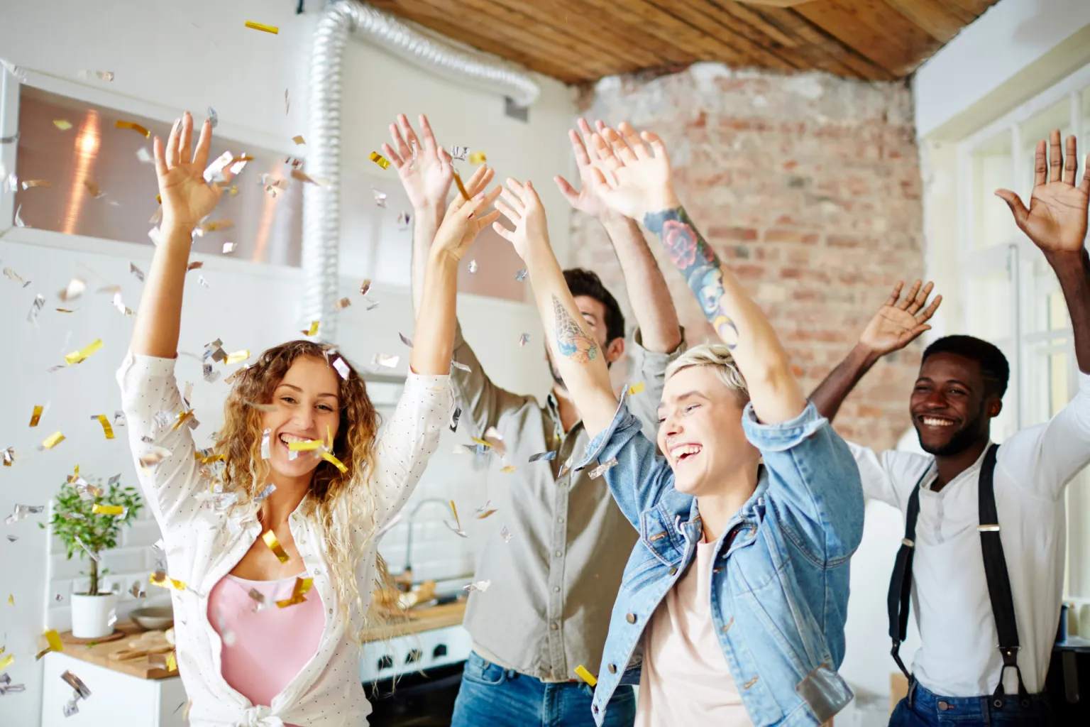 Five friends cheer with their arms raised as gold and silver confetti falls in a bright brick-walled kitchen